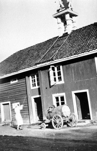 Kvinne ringer i matklokka på stabburet. Vogn med vanntønne utenfor.