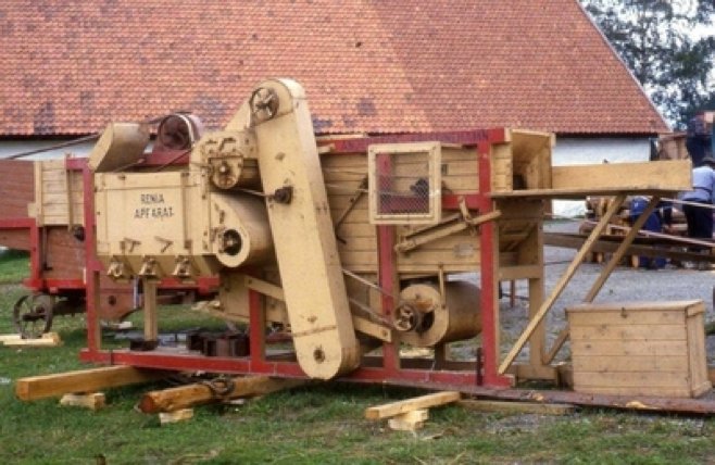 Et trøskeverk av tre med røde rammer, oppstilt foran en låve.